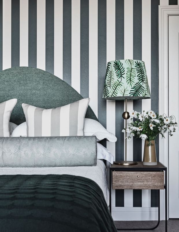 A styled bedroom vignette featuring a bed with a dark green upholstered curved headboard, pale bedding, striped and botanical cushions, and a dark green throw. The room has bold vertical charcoal and white striped wallpaper, a wood bedside table, a brass-toned table lamp with a leafy patterned shade, and a vase of white flowers. White decorative door trim and dark carpet complete the refined, high-contrast scheme.
