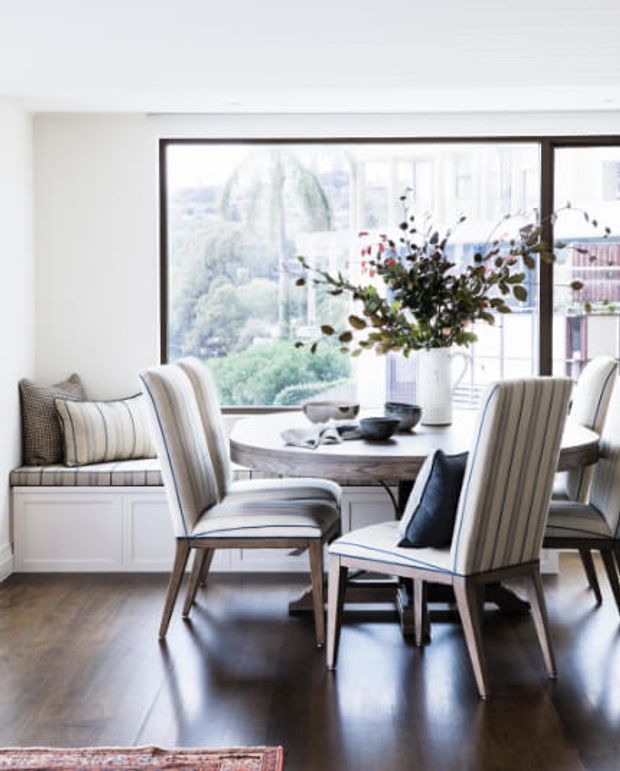 Bright, refined dining area with a round table surrounded by upholstered striped dining chairs, built-in window seating, and a large dark-framed picture window overlooking greenery and nearby buildings. White walls and joinery contrast with dark polished timber flooring, while a central floral arrangement and neutral textiles add a classic, elegant feel.