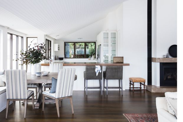 Bright open-plan coastal-style interior combining a kitchen, dining area and living room. The space features dark timber flooring, a white vertically panelled ceiling, white cabinetry with glass-front doors, a timber island benchtop, woven bar stools, and expansive glazed doors and windows. A fireplace with dark surround and timber mantel is visible at right, while the dining area and sofa occupy the foreground.
