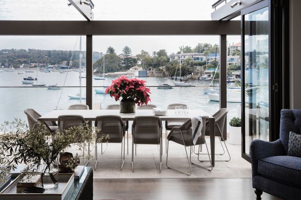 A refined waterfront dining area with a large dining table and woven chairs positioned beside full-height glazed doors and windows. The open glazing frames a marina and sheltered bay, while dark structural framing, pale flooring, indoor plants, and a deep blue armchair create a calm, sophisticated coastal interior.