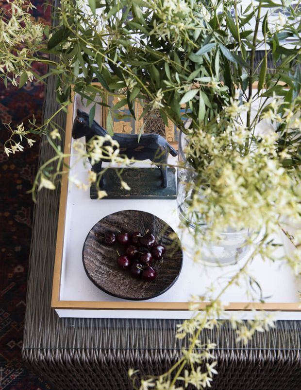 Top-down view of a styled outdoor table or patio setting, partially obscured by leafy flowering branches. The white tabletop has a dark framed edge, a textured dark woven base, a black tray with a glass vessel, and a round dark wooden plate holding cherries. A patterned rug is visible along the left edge.