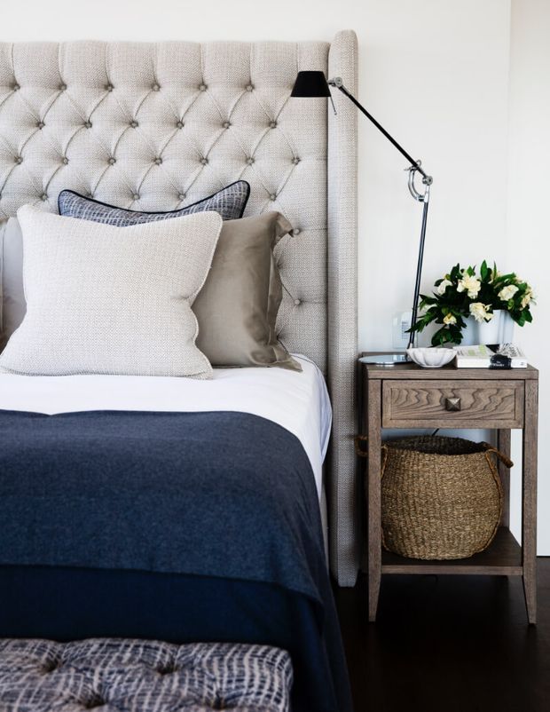 A close-up of a styled bedroom with a tall tufted upholstered bedhead in a light neutral fabric, layered white, beige and navy bedding, and a compact wooden bedside table. A black articulated wall lamp, woven storage basket, and small floral arrangement complete the refined, comfortable setting. Dark timber flooring and a patterned upholstered bench are visible in the foreground.