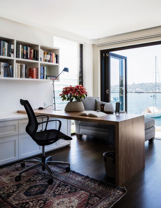 Bright home office overlooking a waterfront, with a large timber desk, ergonomic office chair, built-in white cabinetry and open bookshelves, a grey sofa, dark timber flooring, and full-height glazed doors opening to the view. A patterned rug and potted flowering plant add decorative warmth.