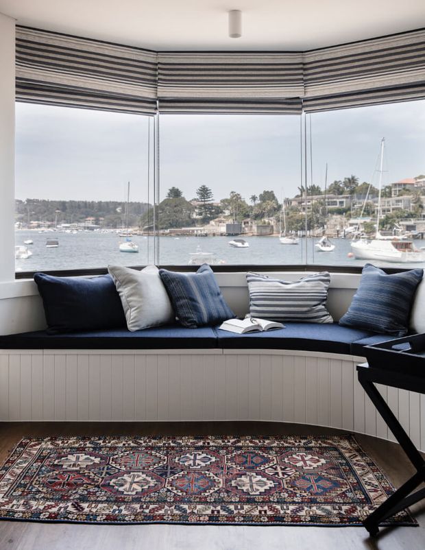 A bright bay-windowed sitting area with a built-in curved window seat upholstered in deep navy, layered with striped cushions and an open book. Full-height glazing overlooks a calm waterfront with boats and homes. Striped Roman blinds frame the windows, while a richly patterned rug adds colour over a light floor. A black keyboard or piano is partially visible at the right.