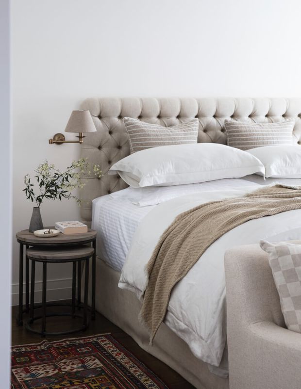 A softly styled bedroom featuring a large tufted upholstered bedhead, layered white bedding, taupe knitted throw, striped cushions, dark wood nesting side tables, and a wall-mounted brass reading lamp. A patterned rug and upholstered armchair add warmth and texture, creating a refined, comfortable bedroom setting.