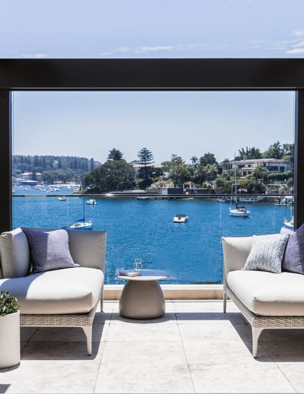 A bright waterfront sitting area framed by large floor-to-ceiling glazing, with two light upholstered lounge chairs, a small round side table, and pale large-format tiled flooring. The view looks across vivid blue water with boats and landscaped residential buildings on the opposite shore.