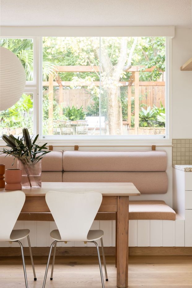 Bright, warmly styled dining nook with a built-in blush upholstered banquette beneath a large picture window. A light timber dining table and white molded chairs sit on pale timber flooring, with white vertical wall panelling, a tiled kitchen edge, indoor plants, and a leafy garden visible outside.