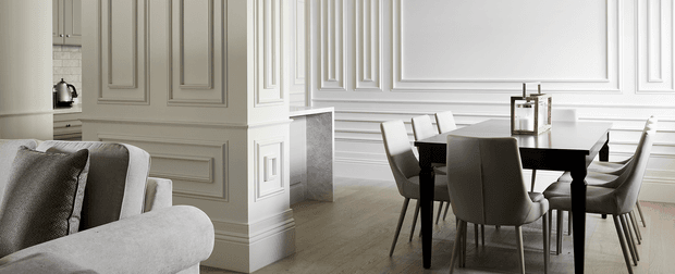 A refined open-plan interior combining a living area and dining space. The room features extensive decorative wall panelling and moulded trim in white, a light timber floor, a dark rectangular dining table with upholstered dining chairs, and a pale upholstered sofa in the foreground. A compact kitchen area is partially visible behind the panelled partition. The overall composition is bright, formal, and restrained.