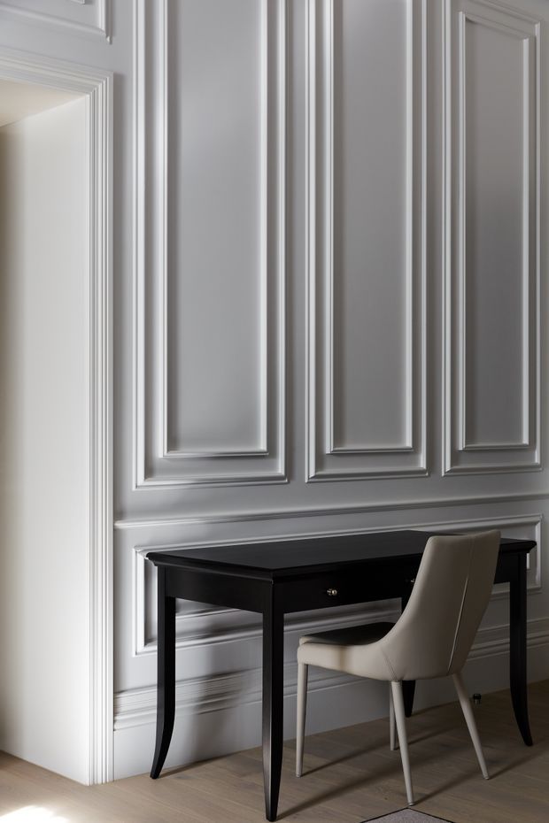 A refined interior vignette showing a compact black writing desk positioned against a grey wall with elaborate raised rectangular moulding and matching decorative door or wall trim. A sculptural cream upholstered chair sits at the desk. Pale timber flooring and soft natural light create a quiet, elegant workspace or console area.