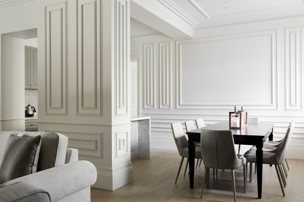 Bright, refined open-plan dining area adjoining a living space and partially visible kitchen. The dining room features a dark rectangular table with upholstered dining chairs, extensive white decorative wall moulding and panelled columns, a framed wall panel, white layered cornices, and light timber flooring. A pale upholstered sofa is visible in the foreground, while the kitchen is glimpsed through an opening at left.