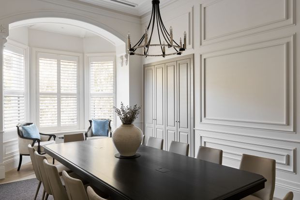 Elegant formal dining room with a large dark rectangular dining table, upholstered dining chairs, white paneled walls and built-in cabinetry, a black lantern-style chandelier, and an arched opening leading to a bright bay-window seating area with plantation shutters. The room has a refined, light-toned classic interior with strong architectural moulding and wainscoting.