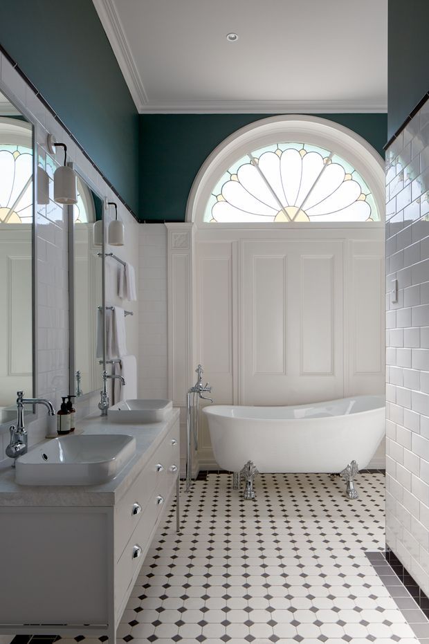 A refined traditional bathroom with a freestanding white clawfoot bathtub, double white vanity, chrome fixtures, and a large mirrored wall. The room features dark teal painted walls, white subway-tile wainscoting, ornate white panelled cabinetry and doors, decorative ceiling cornices, and a distinctive arched fanlight window with stained-glass detailing. The floor is finished in a geometric black-and-white checker pattern.