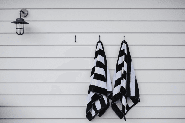 A minimalist exterior wall clad in horizontal white weatherboards, with two black-and-white striped towels hanging from hooks and a small black wall-mounted lantern at the upper left.