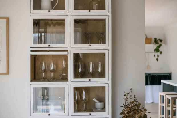 Interior view featuring a tall built-in display cabinet with multiple white-framed glass-front compartments, showcasing glassware, ceramics, and decorative objects. A partial kitchen or dining area is visible through the opening at right, with light timber stools and dark cabinetry. The space has a bright, restrained, contemporary look with warm timber and neutral finishes.