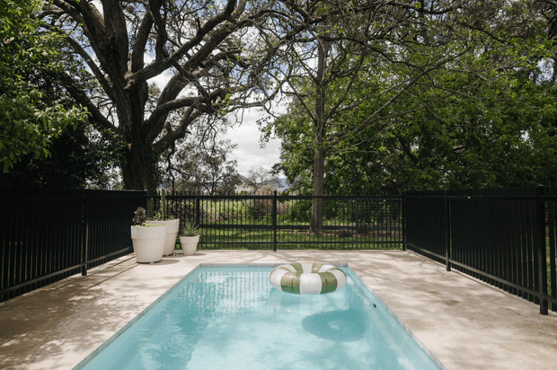 A rectangular swimming pool occupies a paved outdoor terrace, enclosed by black metal fencing and surrounded by mature leafy trees. A floating inflatable ring is visible in the clear blue water, with potted plants positioned along the rear edge. The scene has a private, leafy garden setting and a calm residential outdoor atmosphere.