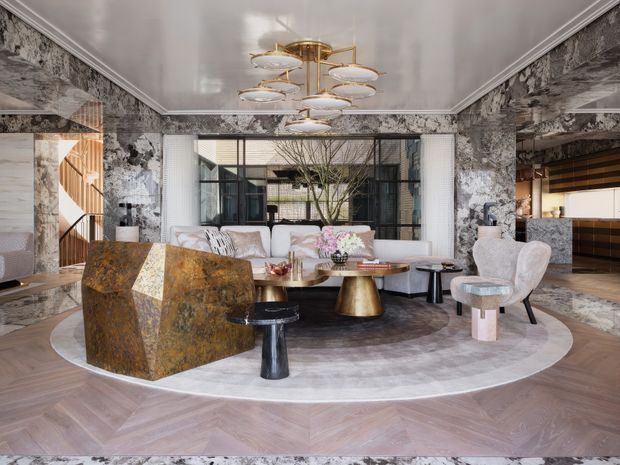 A luxurious, eclectic living room with dramatic marble-patterned walls, herringbone timber flooring, sculptural furniture, a large circular rug, and a striking layered brass-and-glass pendant light. The space includes a white sofa, ornate upholstered chairs, a sculptural stone or metallic lounge chair, a round coffee table, and extensive black-framed glazing opening toward a planted courtyard. A kitchen or bar area is visible through the opening on the right.