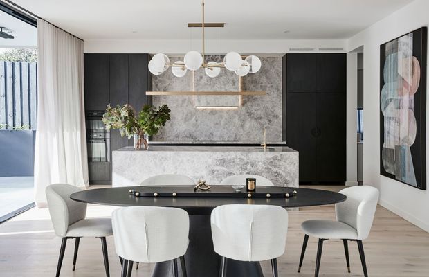 Open-plan dining and kitchen interior with a black oval dining table and upholstered white dining chairs in the foreground. Behind it is a contemporary kitchen featuring dark cabinetry, a large veined stone island and matching splashback, integrated appliances, and a sculptural globe pendant light. Light timber flooring, white walls, sheer curtains, and large glazing create a bright, refined space.