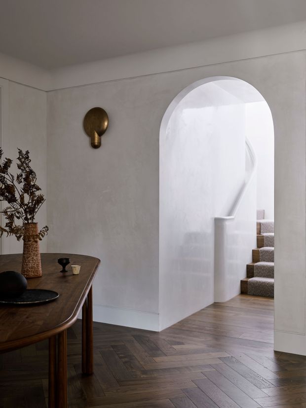 A refined residential entry hall with warm dark timber herringbone flooring, softly textured pale plaster walls, and a large arched opening leading to a carpeted staircase. A dark wooden console table with sculptural decor and dried botanicals sits to the left, while a small brass wall sconce adds a warm accent. The space has a calm, minimalist yet traditional character with curved architectural detailing.
