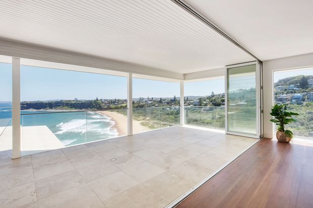 Bright, open-plan glazed living space with full-height corner windows and sliding glass doors overlooking a sandy beach, ocean surf and coastal neighbourhood. The room features large pale stone or porcelain floor tiles, warm timber flooring at the threshold, a white ribbed ceiling, slim light-coloured frames and a potted indoor plant.