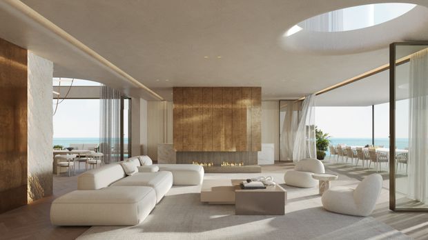 A bright, spacious contemporary living room with a low-profile cream sectional sofa, sculptural lounge chairs, and a simple rectangular coffee table. A large timber-clad feature wall with a linear fireplace anchors the room, while floor-to-ceiling glazing opens toward an ocean view and outdoor dining area. The room has a smooth curved ceiling with a large oval skylight and sheer curtains, creating an airy, refined coastal atmosphere.