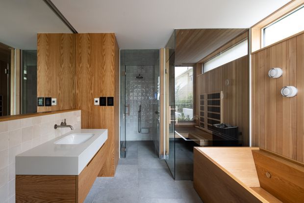 A contemporary bathroom with a long floating timber vanity and white integrated basin, white tiled splashback, large grey floor tiles, and a fully glazed walk-in shower at the centre. Warm wood wall panelling and ceiling lining continue into a separate bathing area with a timber-clad freestanding bathtub. High clerestory windows provide natural light, while dark fixtures and a black towel radiator add contrast.