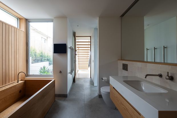 Contemporary bathroom with a freestanding timber bathtub beside a vertical timber slat wall, a large white vanity with timber cabinetry, integrated basin, wall-mounted fixtures and a broad mirror. Grey tiled flooring, pale tiled splashback, and large windows provide natural light. A toilet and hallway are visible beyond the bathroom.