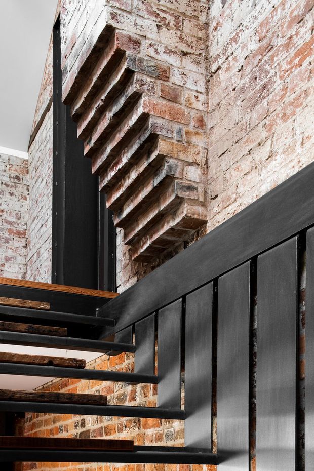 Close-up interior architectural detail of a contemporary industrial stairway. Open-riser stairs combine dark metal stringers, railings and vertical balustrade panels with warm timber treads, set against exposed, weathered red brick walls. The image emphasizes the junction of the stair structure and brick masonry, with strong diagonal lines and textured material contrast.