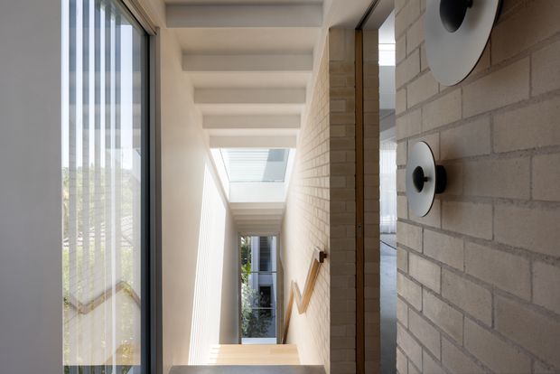 Bright contemporary stairwell and hallway with a long vertical glazed opening on the left, a skylight above the stairs, exposed pale brickwork on the right wall, and minimalist circular wall sconces. A timber handrail runs along the stair wall, with daylight drawing attention toward the upper landing.