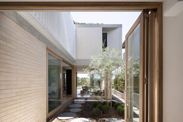 A contemporary courtyard viewed from inside through large timber-framed glazed openings. The enclosed outdoor space features pale brick walls, vertical light-coloured cladding, mature planting, dark gravel or mulch, and circular stepping stones. A covered upper-level connection and flat roof forms create a sheltered, private architectural courtyard.