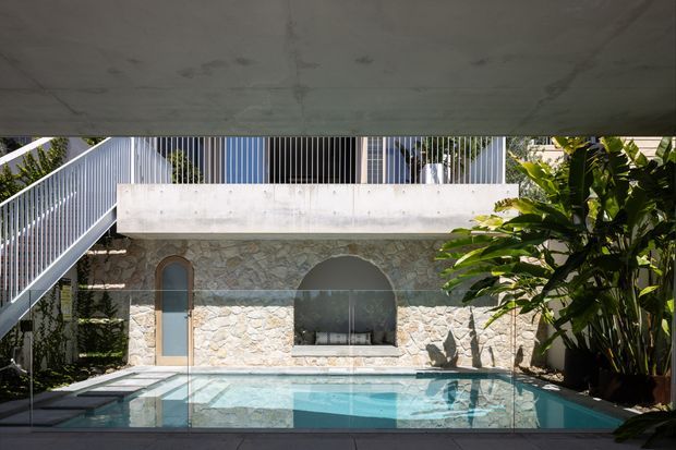 A compact courtyard pool area framed by a concrete overhead slab and surrounded by a stone-clad building. The pool sits directly against a textured pale natural-stone wall with an arched window and narrow arched door, while a white metal stair and balustrade rise along the left side. Lush tropical planting borders the right edge, creating a private, resort-like outdoor setting. The composition shows strong geometric contrasts between the raw concrete, irregular stonework, arched openings, metal railings and reflective turquoise water.