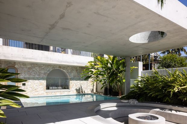 A sunlit modern courtyard pool area beneath a broad cantilevered concrete canopy with a circular roof opening. The space features a pale irregular stone-clad wall with arched niches, a turquoise swimming pool, dark paved flooring, lush tropical planting, and a built-in curved concrete seating or spa-like feature with a round fire-bowl-style element. White and grey architectural finishes create a contemporary resort-like setting.