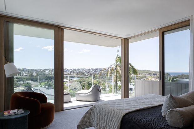 A bright, contemporary bedroom with a low-profile bed, quilted bedding, a sculptural lounge chair, and a floor lamp. Floor-to-ceiling timber-framed glazing wraps around the room, opening to a balcony or terrace with expansive coastal residential views. The space has a restrained, neutral palette and strong indoor-outdoor connection.