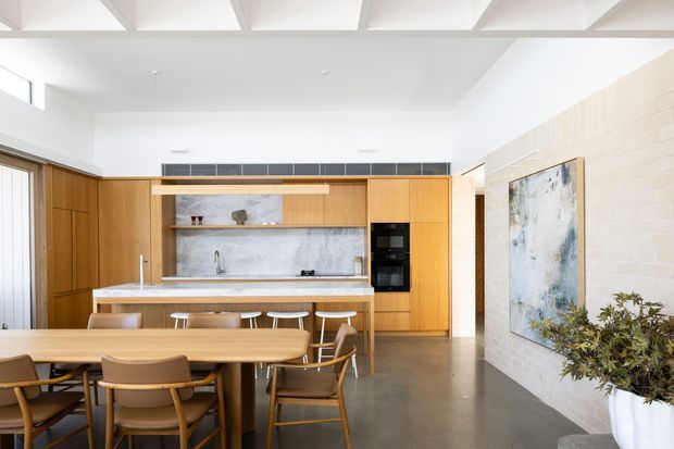 Bright open-plan kitchen and dining area with extensive warm timber cabinetry, a marble-look splashback, central island with stools, and a large timber dining table with chairs. The space features polished concrete flooring, white-painted brick walls, exposed white ceiling beams, skylights, and contemporary pendant lighting. A hallway and artwork are visible beyond the kitchen.