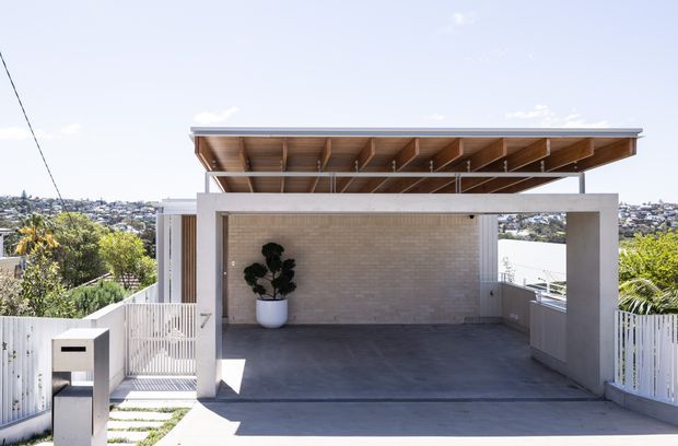 Bright exterior view of a contemporary residential carport or covered outdoor parking area. The structure has a flat, open timber-lined roof with exposed beams, a broad concrete-framed opening, pale brick rear wall, concrete floor, white vertical fencing, and a built-in outdoor bench or storage unit. A suburban hillside neighbourhood is visible beyond the property.
