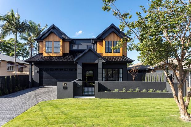 Front exterior view of a contemporary two-storey suburban house with a dark charcoal facade, steep gabled roof forms, warm timber-clad upper-level projections, black-framed windows, an integrated double garage, and a low masonry front wall. The property includes a paved driveway, lawn, boundary fencing, and mature landscaping.