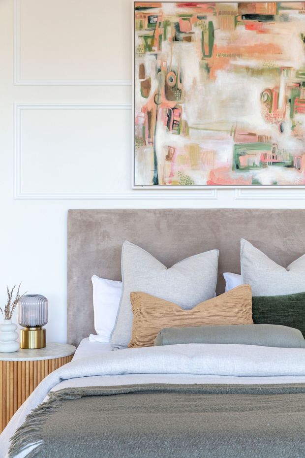 A softly styled bedroom vignette featuring a layered bed with a taupe upholstered headboard, white and pale grey bedding, textured grey throw, and decorative cushions in neutral, ochre, and deep green tones. A round bedside table holds a contemporary lamp and dried botanical arrangement. The wall has white decorative panel moulding and an oversized abstract artwork in muted coral, green, cream, and blush tones.