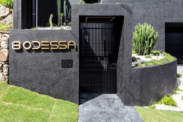 Exterior view of a contemporary dark textured-stucco entrance with a black vertical-slat metal gate, integrated raised planter, sculptural cacti, stone paving and a prominent metallic building sign.