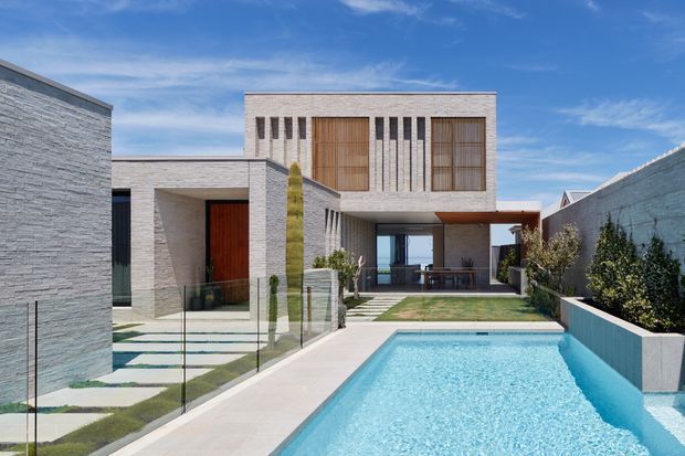 Modern multi-level residential house viewed from the rear, with pale masonry cladding, timber feature doors and screens, tall narrow windows, a covered outdoor living area, landscaped courtyard, and a long rectangular swimming pool in the foreground. The composition emphasizes clean geometric forms, strong horizontal and vertical lines, and a bright blue-sky setting.