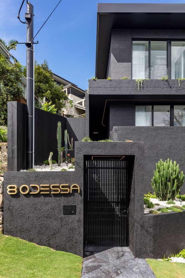 Street-facing view of a contemporary multi-storey residence with a dark textured rendered facade, black metal gate and fencing, projecting box-like balconies, large windows, and landscaped cactus planting. Gold lettering is mounted on the front boundary wall, with a paved entry path and visible overhead utility infrastructure.