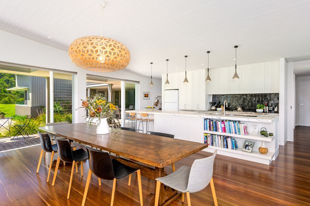 Bright open-plan dining and kitchen interior with a large rustic timber dining table, mixed dining chairs, white built-in kitchen cabinetry, a dark patterned splashback, open shelving with books, pendant lighting, timber flooring, and expansive glazing opening toward a green outdoor area. The space has a warm, contemporary family-home feel.