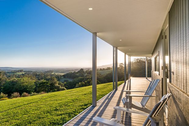 A covered verandah or balcony terrace runs along the side of a contemporary home, with slim posts, a slatted ceiling, timber decking, and outdoor lounge chairs. The terrace overlooks a broad green rural landscape and rolling hills in warm late-afternoon light. The building facade combines vertical timber cladding with a textured stone or concrete lower wall.