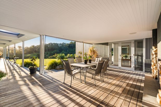 A spacious covered outdoor deck or terrace adjoining a contemporary home, furnished with a rectangular dining table and several outdoor chairs. Timber decking, slender structural posts, a white-lined ceiling, glazed sliding doors, and dark vertical exterior cladding frame views toward a landscaped lawn and trees. Strong afternoon sunlight creates long shadows across the deck, giving the space an open and inviting entertaining atmosphere.