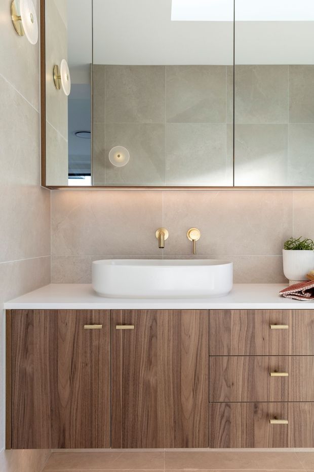 A refined contemporary bathroom vanity featuring a white vessel basin on a white benchtop, wall-mounted brass tapware, warm beige stone-look wall and floor tiles, and dark walnut-toned timber cabinetry with brass handles. A large mirrored cabinet spans the wall above the vanity, with soft integrated under-cabinet lighting and a skylight or high window providing natural light.