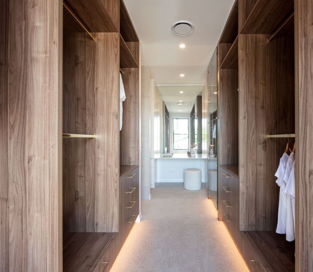 A long, narrow walk-in wardrobe with full-height dark timber built-in cabinetry, open shelving, drawers, hanging rails and integrated warm under-cabinet lighting. The wardrobe leads directly toward a bright bathroom vanity area visible at the far end, with a mirror, basin and freestanding cylindrical stool or ottoman. Neutral carpet, white ceiling and recessed lighting create a refined, warm, contemporary interior.