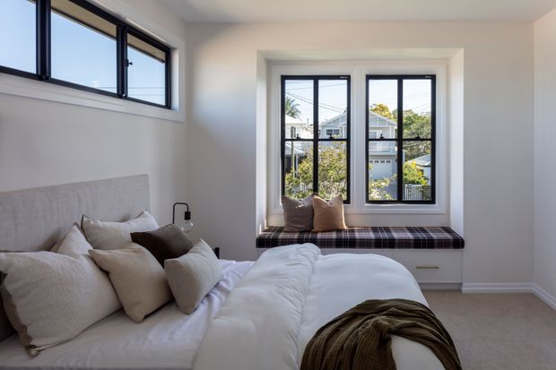 Bright, compact bedroom with a low-profile upholstered bed, layered neutral bedding and cushions, and a built-in window seat beneath large black-framed windows. The room has pale walls, light carpet, and abundant daylight, with an urban residential view beyond the glazing.