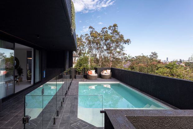 Contemporary rooftop-style terrace with a rectangular swimming pool beside a dark-clad modern residence. Frameless glass balustrades line the pool edge, with dark tiled paving, built-in seating, and landscaped suburban views beyond.