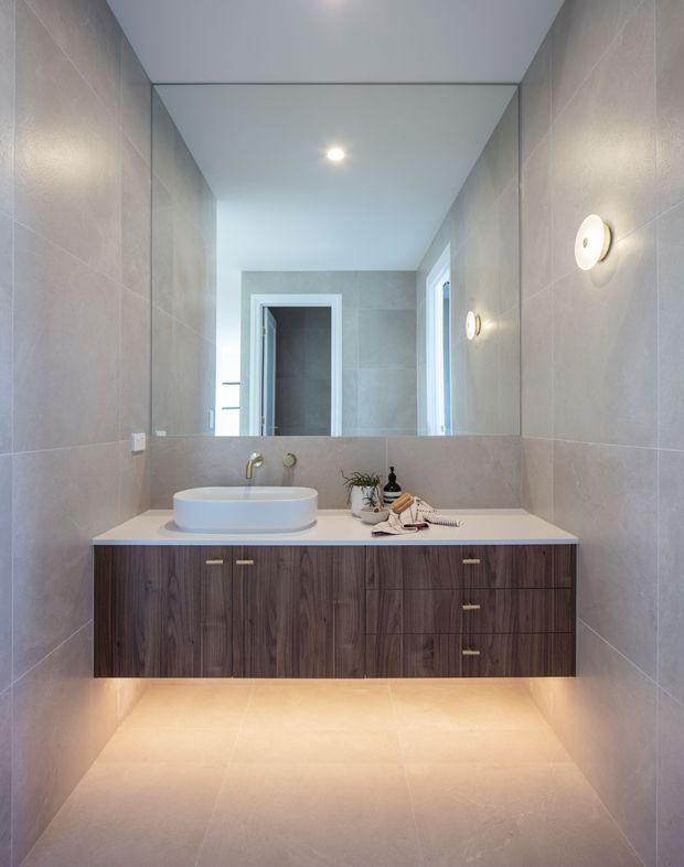 Contemporary bathroom vanity with a floating dark timber-grain cabinet, white vessel basin, pale stone-look wall and floor tiles, large wall-to-wall mirror, and warm under-cabinet lighting. The compact room has clean lines, recessed ceiling lighting, and minimal styling.