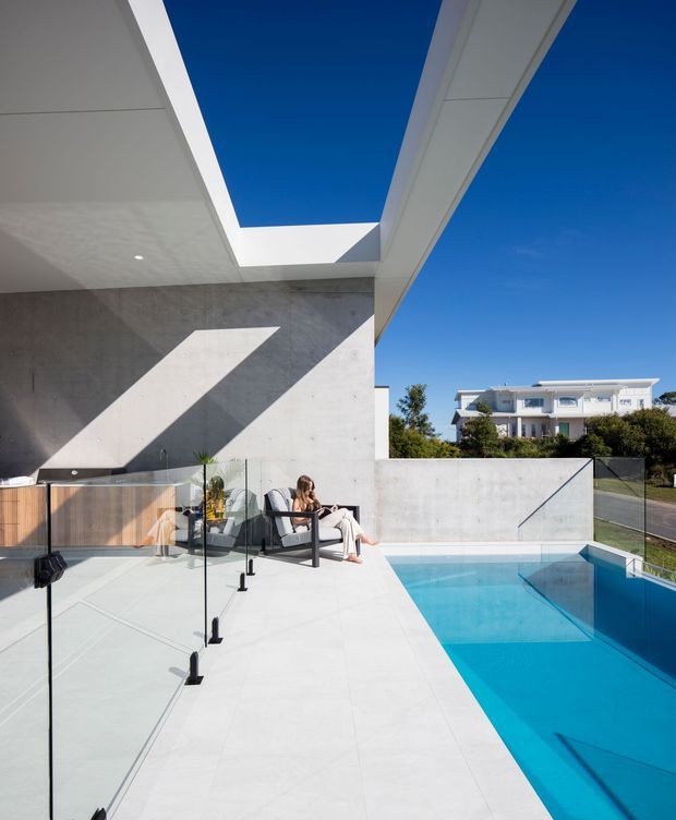 Contemporary outdoor pool terrace with a crisp white tiled deck, frameless glass balustrades, a turquoise swimming pool, and a dramatic concrete wall beneath a large rectangular roof opening. Strong geometric shadows and clean modern lines define the scene, with a seated person and neighboring homes visible in the background.