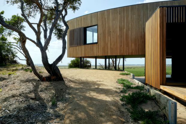 Exterior view of a contemporary elevated timber-clad house set lightly into a rural or coastal landscape. The building has a long rectangular upper volume raised on slender dark supports, vertical timber cladding, a projecting entrance volume with deep vertical screening, and large openings. Native trees, sandy ground, and open fields or water beyond reinforce the secluded site setting.