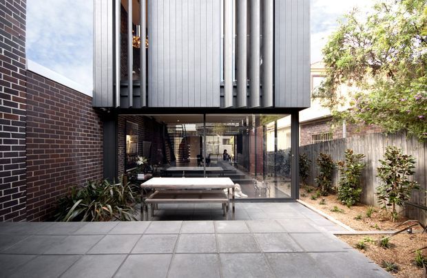 Contemporary two-storey extension viewed from a paved courtyard. A dark brick wall frames a glazed ground-floor living area, while the upper level projects overhead with vertical silver-grey metal cladding and slender screening fins. The courtyard includes a timber dining table, planting beds, and a timber boundary fence.